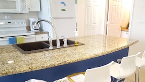 A kitchen with a granite countertop, stainless steel sink, and white appliances. There are three white chairs and a blue wall. - Waterfront Condo Family Getaway (St Petersburg)