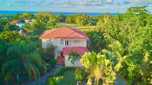 A two-story house with a red roof, surrounded by lush greenery and a view of the ocean. - Spacious 5 Bedroom Villa, Centrally Located, Near Beach. (Runaway Bay)
