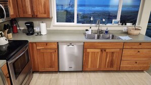 A modern kitchen with wooden cabinets, a stainless steel dishwasher, a double sink, and a large window overlooking the ocean. - Ocean Front ON the Beach - Spectacular Views! 14 Minutes to Bandon Dunes Golf! (Bandon)