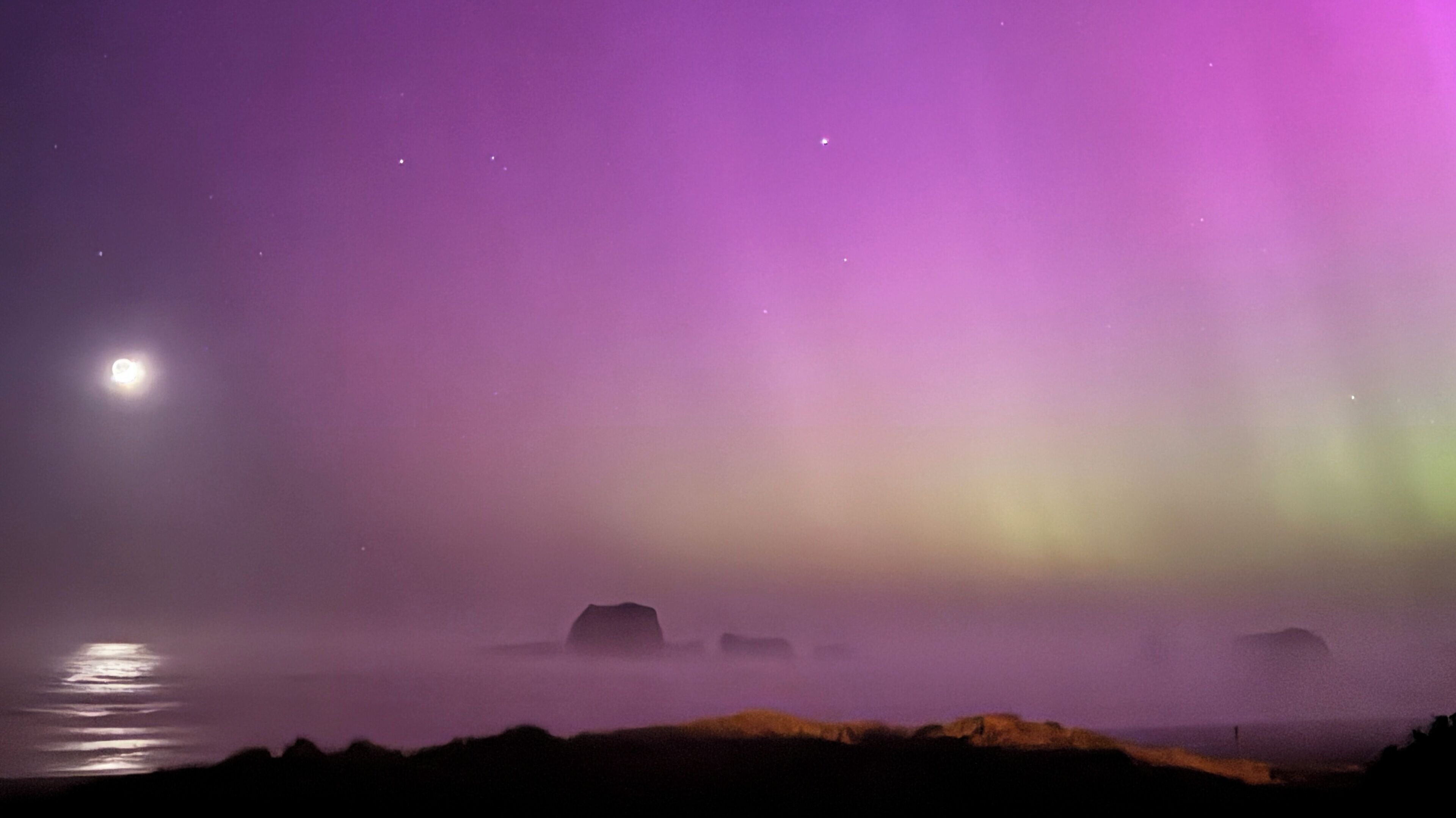 A night scene with a visible moon and aurora borealis over a rocky coastline.