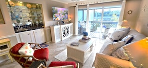 A living room with a sofa, a chair, a coffee table, and a television. There is a balcony with a view of the outdoors.