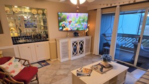 Ein Wohnzimmer mit Fernseher, einer Bar inklusive Gläsern und einem Balkon mit Gartenmöbeln.