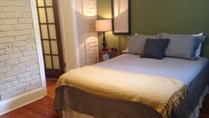 A bedroom with a wooden floor, a bed with a beige and brown bedspread, a nightstand with a lamp, and a framed picture on the wall. - The Garden Apartment with a Murphy Table (Savannah)