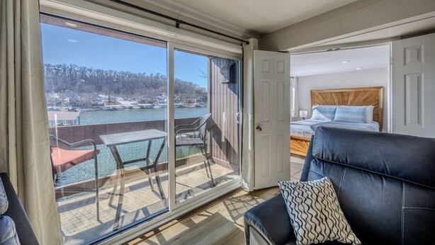 A balcony with a table and chairs, a bed with a wooden headboard, and a view of a body of water and trees. - 22 Mile Marker! Indoor/Outdoor Pools, Hot Tub! New (Osage Beach)