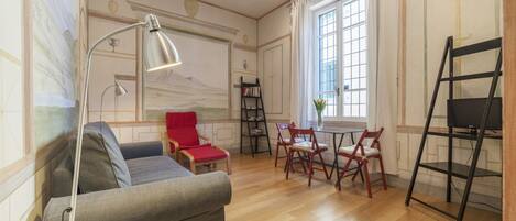 Un salon comprenant un canapé, un fauteuil rouge, une table avec des chaises, une lampe, une bibliothèque et un téléviseur posé sur un meuble.