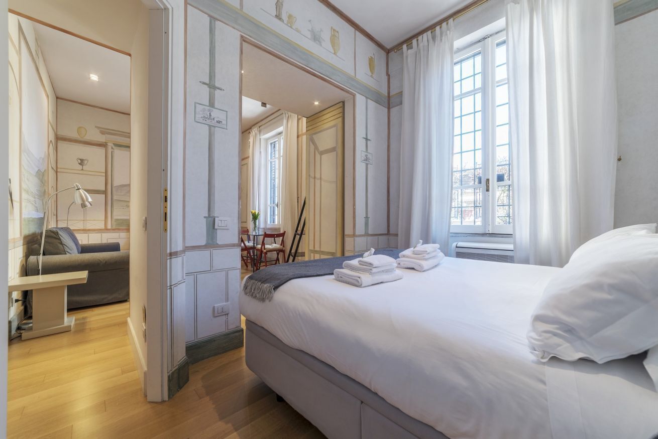 A hotel room with a large bed, a desk, and a chair. There is a window with curtains, a door, and a mirror on the wall.