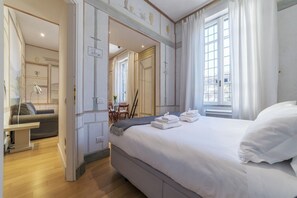 Habitación de hotel con una cama grande, un escritorio y una silla. Hay una ventana con cortinas, una puerta y un espejo en la pared.