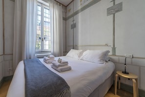 A neatly made bed with white linens and a grey throw blanket, a bedside table with a lamp, and a window with curtains.