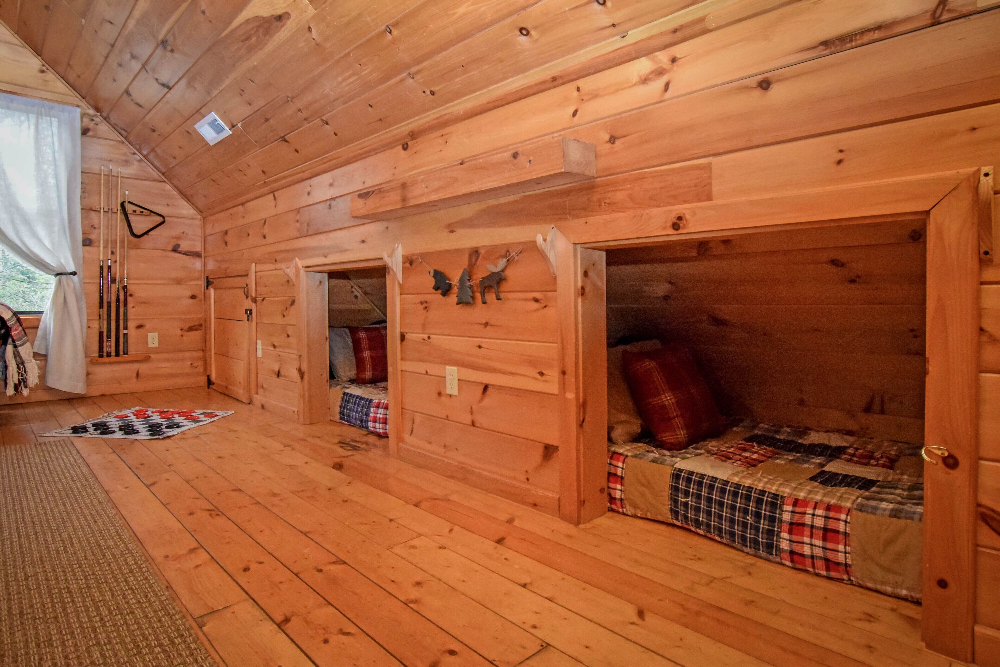 Une cabane aux murs et au plancher en bois, un lit en mezzanine avec une couverture à carreaux, une petite table avec une chaise et une fenêtre avec des rideaux.