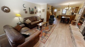 A living room with a brown leather sofa, a coffee table, and a patterned rug. There is a dining area with a wooden table and chairs. The room has a kitchen area with a stone backsplash and a chandelier.