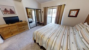 A bedroom with a bed, a wooden dresser, a television, a mirror, and a sliding glass door leading to a balcony.