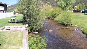 Un arroyo cristalino con un banco de madera y una mesa de picnic, rodeado de vegetación y casas al fondo.
