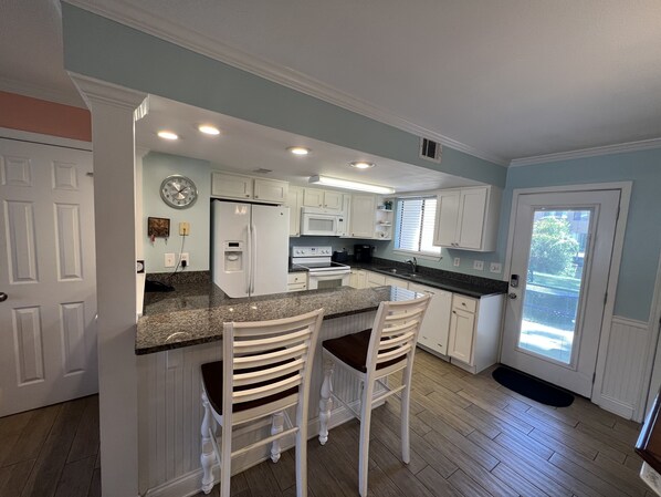 A kitchen with white cabinets, a granite countertop, a refrigerator, a microwave, and two bar stools.