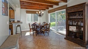 A dining area with a wooden ceiling, a chandelier, a dining table with chairs, a kitchen area, and a glass cabinet with decorative items.