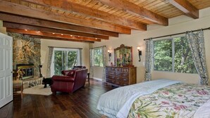 A spacious bedroom with a large bed, wooden beams on the ceiling, a stone fireplace, and large windows with curtains.