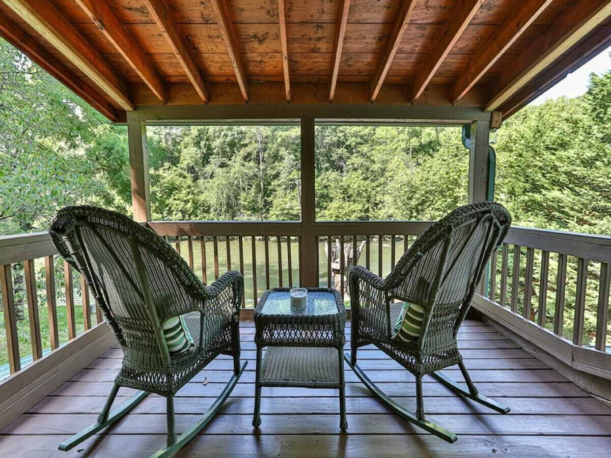 Un espace extérieur abrité, doté de deux fauteuils en osier et d’une petite table, entouré d’une terrasse en bois et d’une rambarde, et offrant une vue sur une zone boisée.