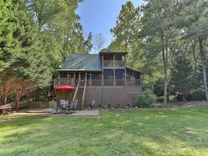 Une cabane à deux étages avec un balcon, entourée d’une cour verdoyante et d’arbres.