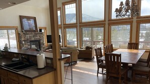 A spacious kitchen with a stone fireplace, large windows, and a dining area with wooden furniture. - Spacious Family Home In Town With Spectacular Lake And Mountain Views (Grand Lake)