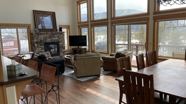 A spacious living room with a fireplace, large windows, and a dining area. - Spacious Family Home In Town With Spectacular Lake And Mountain Views (Grand Lake)