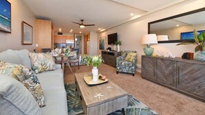 A spacious living area with a sofa, coffee table, and a dining table with chairs. There is a kitchen in the background with cabinets and a refrigerator. - Amazing Views, Direct Oceanfront Studio w Pool AC - Maui Kai 206 (Lahaina)