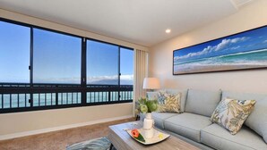 A room with a large window overlooking the ocean, a sofa with patterned cushions, a coffee table with decorative items, and a painting of a beach scene. - Amazing Views, Direct Oceanfront Studio w Pool AC - Maui Kai 206 (Lahaina)