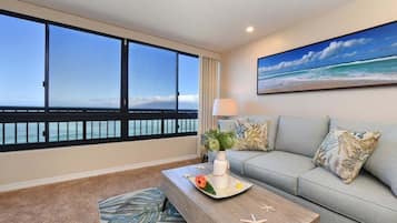 Een kamer met een groot raam dat uitkijkt op de oceaan, een bank met gestreepte kussens, een bijzettafeltje met decoratieve objecten, en een schilderij van een strandgezicht.