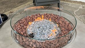Un foyer extérieur en verre, doté d’un cadre métallique, entouré de gravier, installé sur une terrasse en béton.