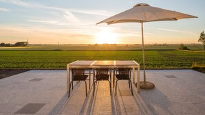 Une table entourée de chaises et protégée par un parasol, posée sur une surface en béton, d’où l’on peut admirer un champ verdoyant avec un coucher de soleil en arrière-plan.