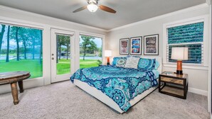 A bedroom with a large bed, a ceiling fan, a nightstand, and a view of a green landscape through the door.