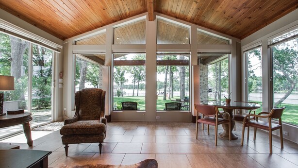 A spacious room with a high ceiling, large windows, a dining area with a round table and chairs, and a comfortable armchair.