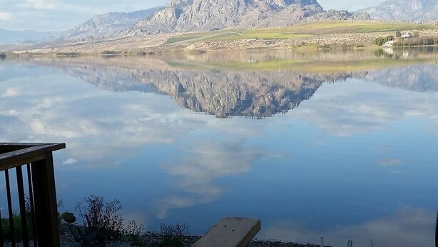 A lakeside view with a wooden deck and chairs, a mountain in the background, and a clear sky.