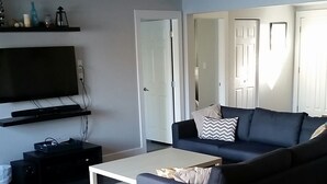A modern living room with a grey sofa, a coffee table, and a TV mounted on the wall. There is a door leading to another room and a small table with a lamp.