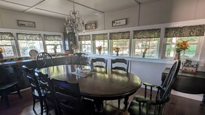Uma sala de jantar com uma mesa redonda rodeada de cadeiras, grandes janelas com cortinas decorativas e um lustre.