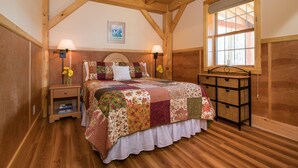 Een slaapkamer met een houten plafond, een groot bed met een bloemenquilt, twee bedlampen, een nachtkastje met boeken en een raam met uitzicht.