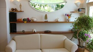 A living room with a beige sofa, wooden shelves with decorative items, a potted plant, and a circular window with a view of greenery. - great sea view - great position - not far from the town center (Positano)
