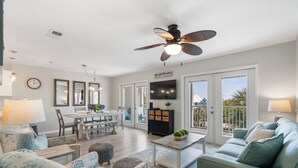 A spacious living room with a dining area, a ceiling fan, a TV, and a balcony with a view. - Gulf Place Caribbean - Top Rated Condo on 30-A with Pickleball & Tennis! (Santa Rosa Beach)