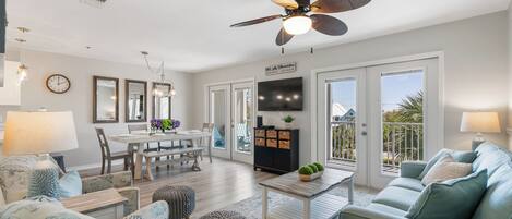 A spacious living room with a dining area, a ceiling fan, a TV, and a balcony with a view.