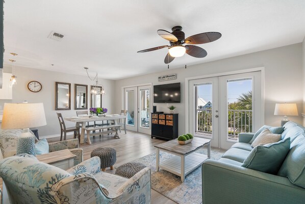 A spacious living room with a dining area, a ceiling fan, a TV, and a balcony with a view.