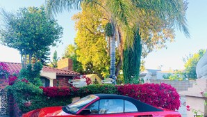 A red convertible car parked in a driveway with a brick house, lush greenery, and a statue in the background. - Beautiful Spanish style home- Garden. Fremont, California.  (Fremont)