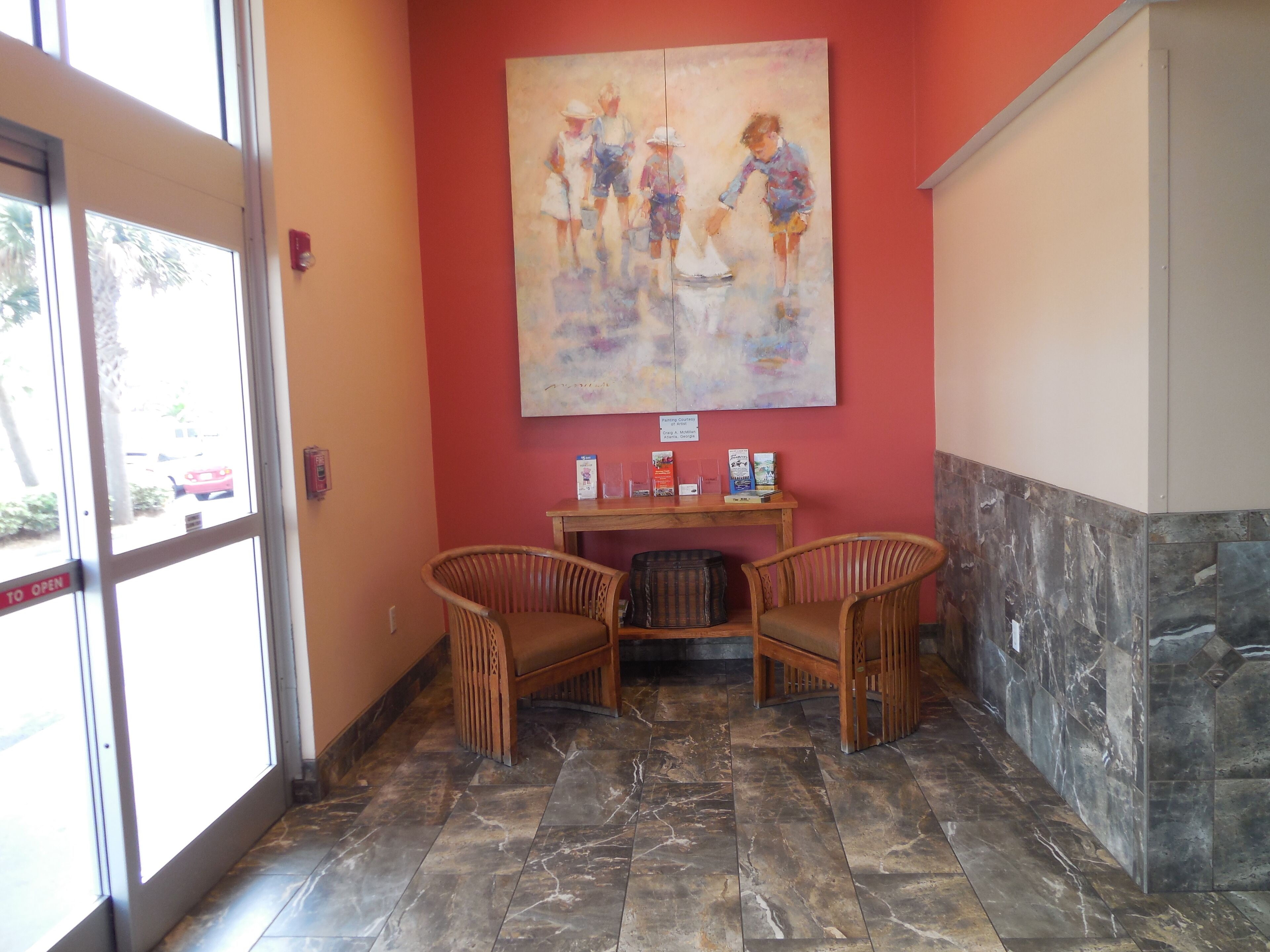 A reception area with a painting, two wicker chairs, a small table with brochures, and a stone-tiled floor.