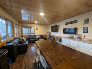 Acogedor interior de cabaña con mesa de comedor de madera, sofá de cuero y zona de cocina con televisor y estación de café.
