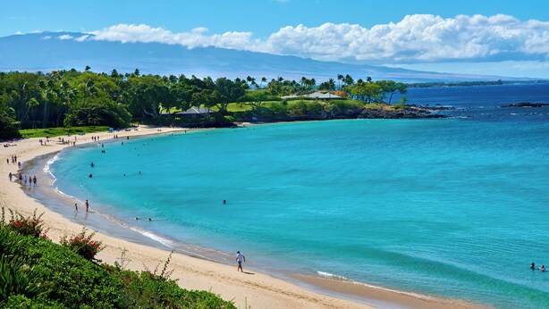 Une plage à l’eau turquoise et limpide, où l’on peut voir des gens nager et se promener le long du rivage, avec un paysage verdoyant luxuriant en arrière-plan.