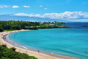 A beach with clear turquoise water, people swimming and walking along the shore, and a lush green landscape in the background.