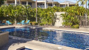 Une piscine entourée de chaises longues et de palmiers, le tout bordé d’un immeuble doté de nombreux balcons.