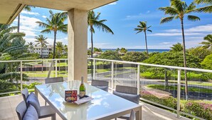 Un balcon avec une table en verre, des chaises et une bouteille de champagne. Il donne sur une route bordée de palmiers, avec un bâtiment visible au loin.