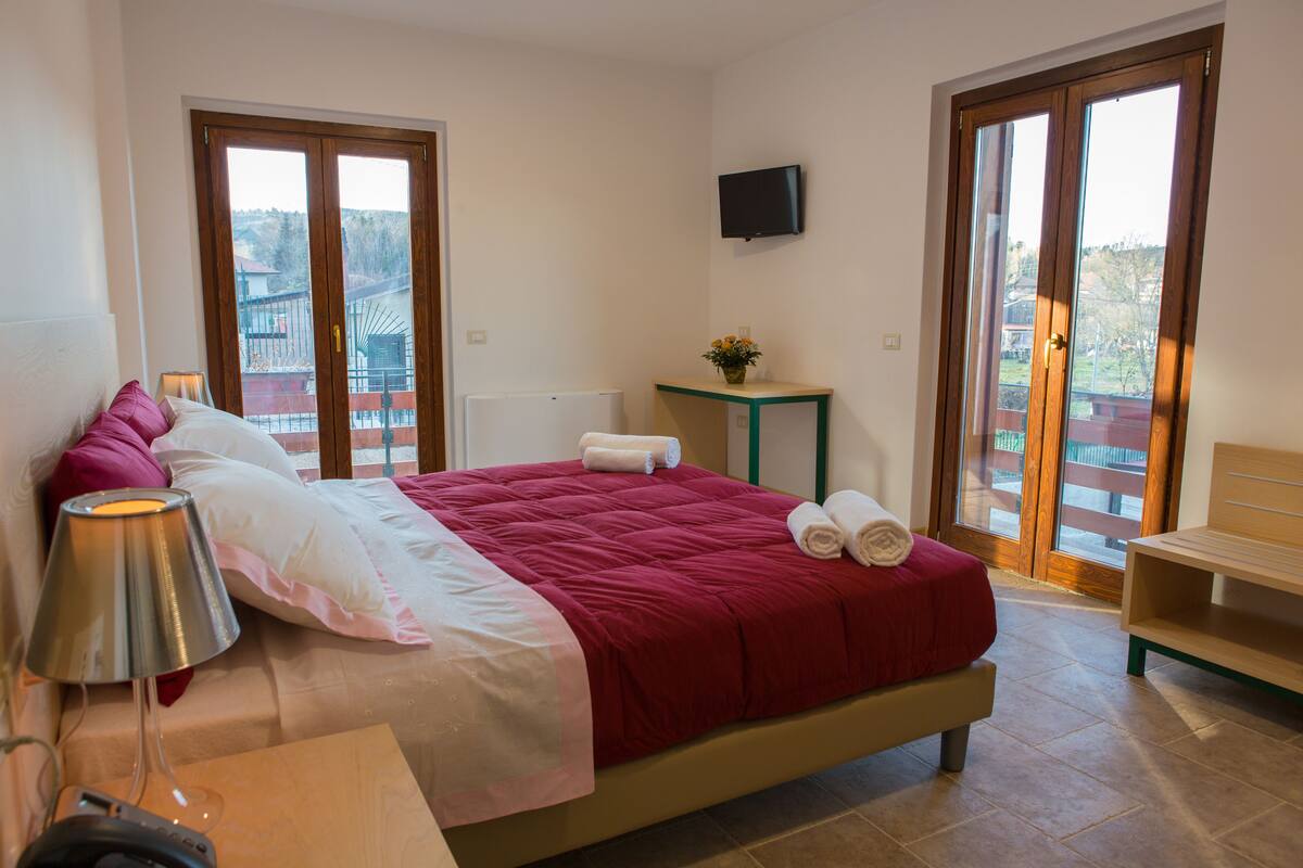 Un dormitorio con cama, una mesita de noche con lámpara, una pequeña mesa con una flor y un televisor en la pared.