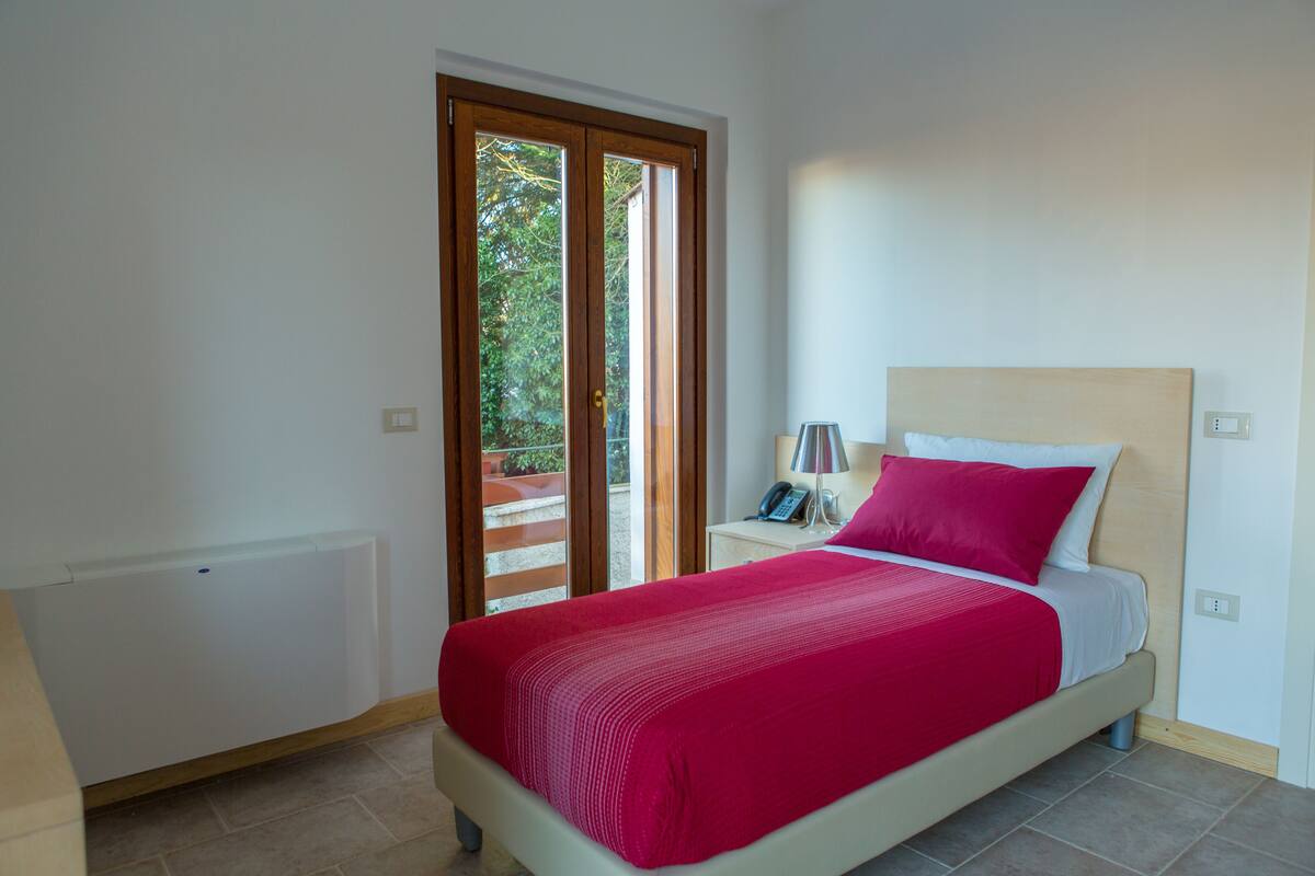 Un dormitorio con cama, mesita de noche, lámpara, radiador y puerta corrediza de vidrio que da a un área exterior.
