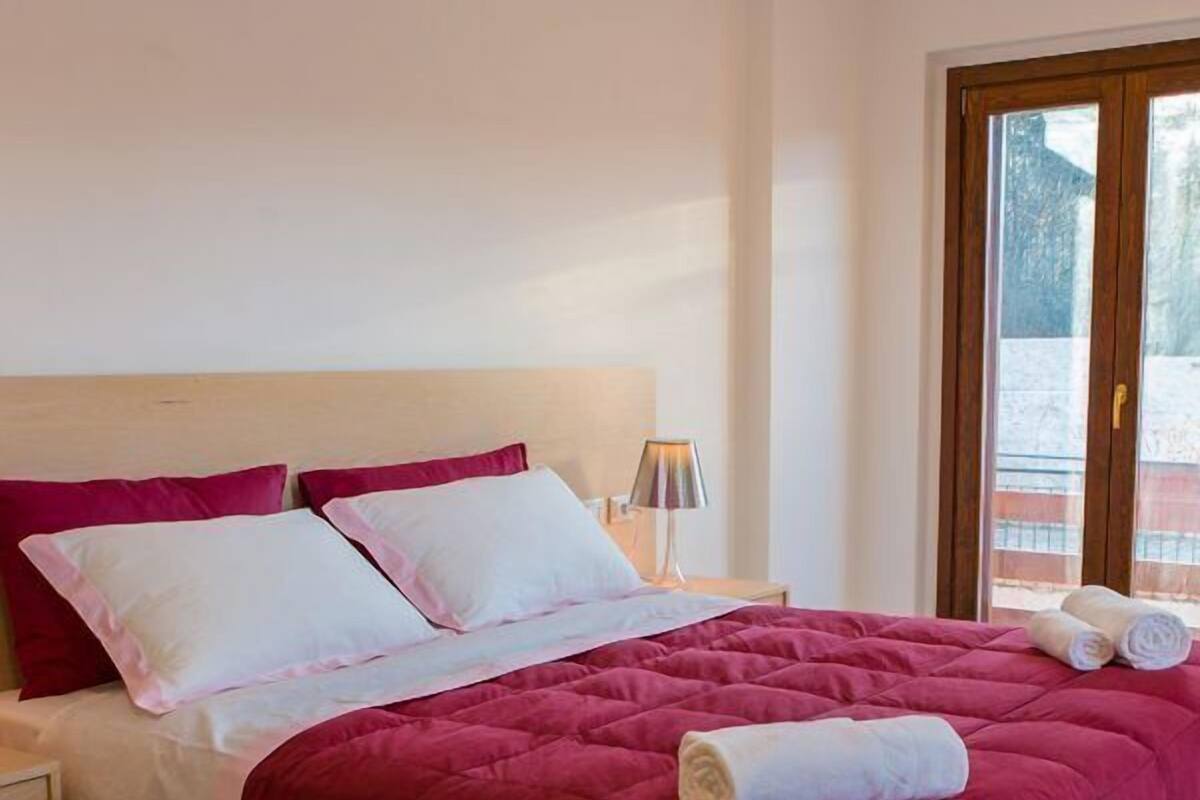 Una cama bien hecha con un edredón rojo, almohadas blancas y una toalla enrollada al lado.