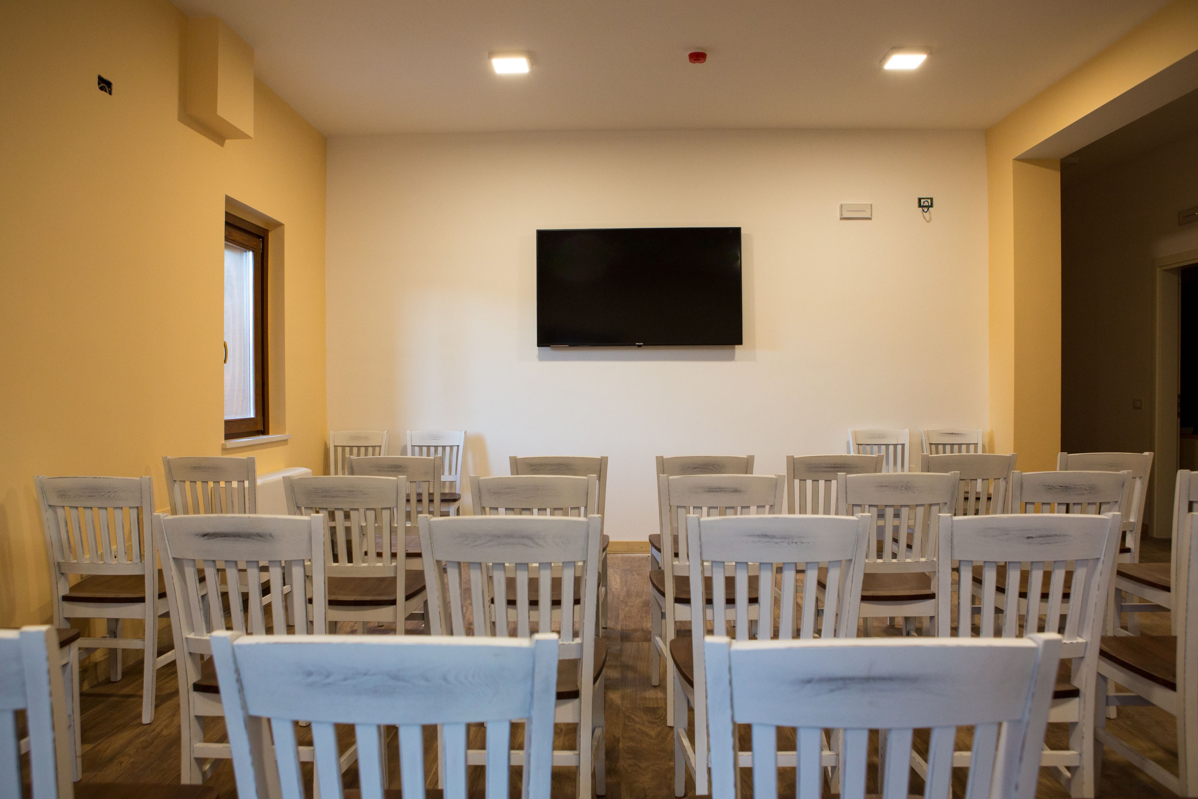 A conference room with white chairs arranged in rows, a flat-screen TV mounted on the wall, and a window on the left side.