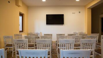A conference room with white chairs arranged in rows, a flat-screen TV mounted on the wall, and a window on the left side.